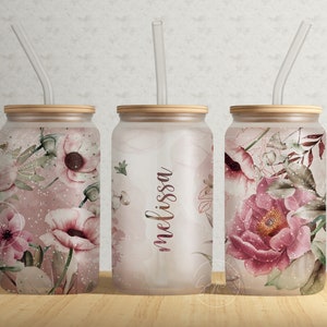 Puede incluir: Tres vasos de vidrio transparente con tapas de bambú y pajitas a juego. Cada vaso presenta un diseño floral diferente con flores rosas y blancas y hojas verdes. El vaso del medio tiene el nombre "melissa" escrito en él.