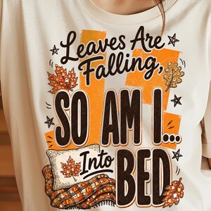 May include: A cream-coloured t-shirt featuring a graphic print with the text "Leaves Are Falling, SO AM I... Into BED". The design includes orange rectangular accents, autumn leaves, a white pillow, a plaid blanket, and small black stars.