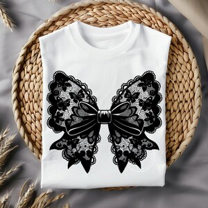 May include: A white t-shirt with a black lace bow design. The bow is made of intricate lace with a floral pattern and a center bow.