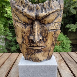 Peut inclure: Une sculpture en bronze texturé représentant un visage, montée sur une base carrée en marbre gris. Le visage affiche une expression sévère. Le fond est vert et flou. La surface de la sculpture est rugueuse et vieillie.