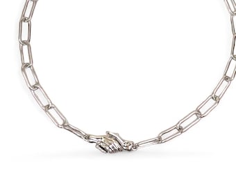 May include: Silver-toned chain necklace with a unique design. The necklace features a series of elongated rectangular links. A detailed hand clasp is at the center. The necklace has a lobster clasp closure and an adjustable chain. The necklace is displayed on a white background.
