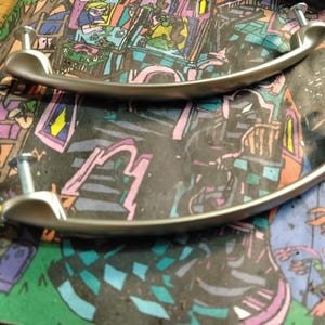 Puede incluir: Dos tiradores de armario plateados con diseño curvo. Cada tirador tiene dos tornillos para el montaje. Los tiradores están colocados sobre una superficie colorida y estampada con ilustraciones de dibujos animados.