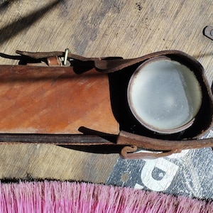 Könnte beinhalten: Ein braunes Lederetui mit einem Riemen und einer runden Glaslinse. Das Etui ist für eine kleine Taschenlampe oder ein anderes Werkzeug gedacht.