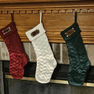 May include: Three cable-knit Christmas stockings in red, white, and dark green. Each stocking features a folded cuff with a brown leather tag attached by two metal rivets. The tags are engraved with the names MOM, SARA, and DAD respectively.