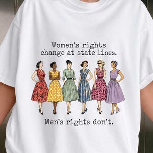 以下が含まれることがあります： ヴィンテージスタイルのドレスを着た6人のイラストが描かれた白いTシャツ。上部には「Women's rights change at state lines.」、下部には「Men's rights don't.」というテキストがあります。ドレスは赤、黄、緑、青、ピンク、紫の色合いです。