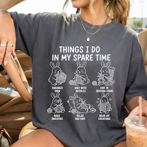May include: Dark gray t-shirt with the text "THINGS I DO IN MY SPARE TIME" and illustrations of rabbits knitting and organizing yarn. The shirt also has the phrases "ORGANIZE YARN", "KNIT WITH NEEDLES", "KNIT IN ROCKING CHAIR", "MAKE SWEATERS", "RELAX AND KNIT", and "WEAR MY CREATIONS".