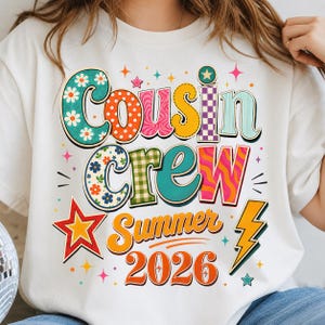 May include: A white t-shirt featuring a colorful graphic with the text Cousin Crew Summer 2026. The design uses various patterns including flowers, polka dots, and checkers in shades of orange, teal, yellow, and pink, accented by stars and a lightning bolt.