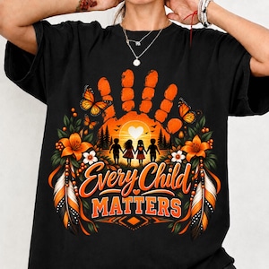 May include: A black t-shirt featuring a graphic of an orange handprint containing a sunset scene with four children holding hands, butterflies, and orange flowers. The text "Every Child MATTERS" is printed in orange below the graphic.