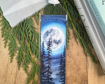 May include: A rectangular bookmark featuring a watercolor painting of a full moon glowing above a dark forest of pine trees. The bookmark is encased in a clear plastic sleeve and is topped with a frayed white fabric ribbon.