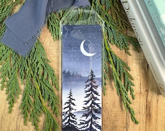 May include: A rectangular bookmark encased in a clear plastic sleeve, featuring a watercolor illustration of a night landscape with pine trees under a crescent moon and stars, topped with a frayed slate blue fabric ribbon.