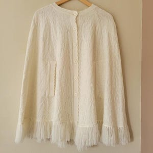 Peut inclure: Cape en tricot crème avec un devant boutonné et un bord festonné. La cape présente un motif texturé et une bordure à franges en bas. Elle a une encolure ronde et une poche sur le côté gauche.