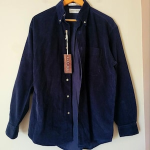 Puede incluir: Una camisa de pana azul marino con mangas largas y botones. La camisa tiene un bolsillo en el pecho y una etiqueta colgante que dice "RUSTIQUE". La camisa está colgada en una percha de madera.