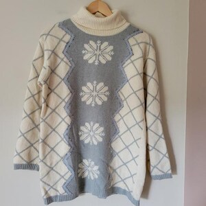 Pode incluir: Camisola creme de gola alta com um desenho de floco de neve. A camisola apresenta um padrão de losangos azul claro e branco nas mangas e laterais, com um painel central azul claro adornado com flocos de neve brancos. A camisola está pendurada num cabide de madeira.