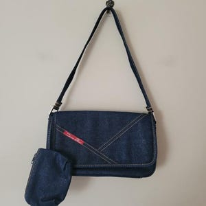 May include: A dark blue denim shoulder bag with a matching small pouch. The bag features white stitching and a flap closure. A red label with white text is on the front. The strap is long.