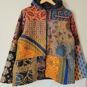 May include: A quilted zip-up hooded jacket featuring a patchwork design with various colourful patterns including floral, geometric, and abstract motifs in shades of orange, blue, black, and brown. The jacket has a solid black hood and a front zipper.
