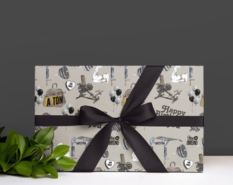 Puede incluir: Un regalo envuelto con un lazo negro. El papel de regalo tiene un fondo beige con ilustraciones de equipos de gimnasio, globos y el texto "I love you a ton" y "Happy Birthday".