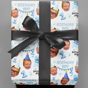 May include: A rectangular gift box wrapped in white paper featuring a repeating pattern of a baby's face, blue party hats, the number 1, and the text Birthday Boy, tied with a wide black satin ribbon centered in a bow across the front.