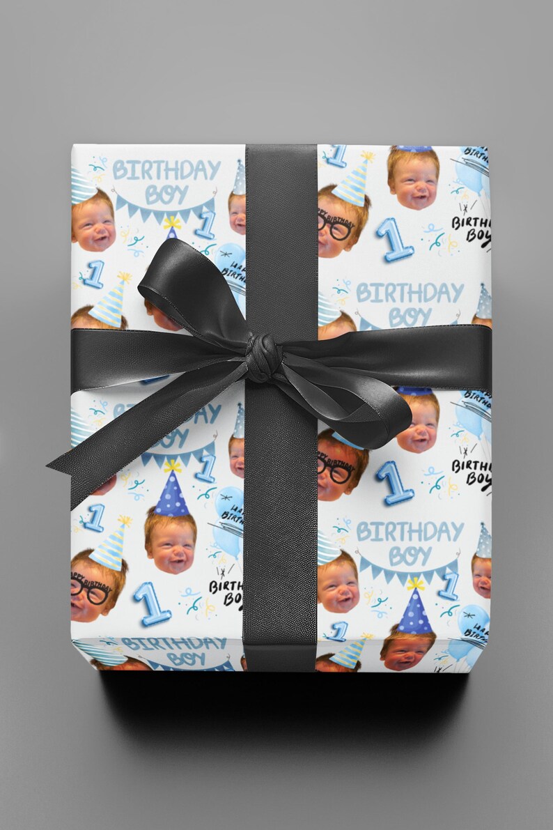 May include: A rectangular gift box wrapped in white paper featuring a repeating pattern of a baby's face, blue party hats, the number 1, and the text Birthday Boy, tied with a wide black satin ribbon centered in a bow across the front.