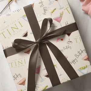 May include: A white gift box with a brown ribbon wrapped around it. The box is decorated with a pattern of martini glasses and the text "You're just a Tini bit older" and "Happy Birthday".