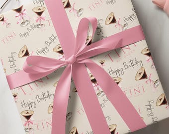Op de afbeelding: Wit cadeaupapier met een roze lint. Het papier heeft een patroon van martini glazen met de tekst "TINI BIT OLDER" en "Happy Birthday".