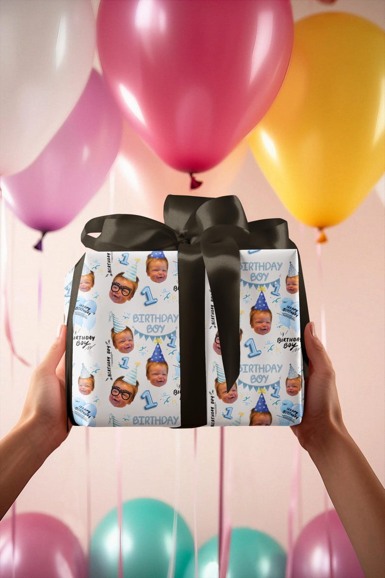 May include: A square gift box wrapped in white paper featuring a repeating pattern of a baby's face wearing a blue party hat, the text "BIRTHDAY BOY", and the number "1" in blue, topped with a large, tied black satin ribbon bow.