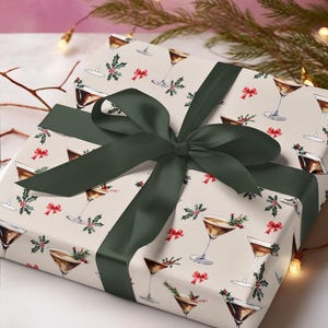K&ouml;nnte beinhalten: Eine wei&szlig;e Geschenkbox mit einem gr&uuml;nen Band und einem Muster aus braunen Martini-Gl&auml;sern mit Stechpalme und roten Schleifen.