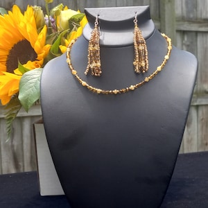 Puede incluir: Un collar dorado con un colgante de borla de cuentas marrones y doradas. El collar se muestra en un maniquí de busto negro.