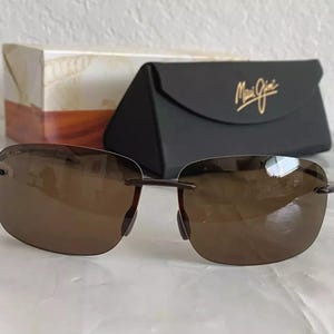 May include: Brown sunglasses with a metal frame and brown lenses. The sunglasses are sitting on a table in front of a black case with the text "Maui Jim" on it.