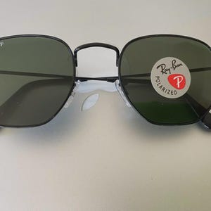 May include: Black octagonal sunglasses with green lenses. The sunglasses have a black frame and the Ray-Ban logo is visible on the lens. The lenses are polarized.