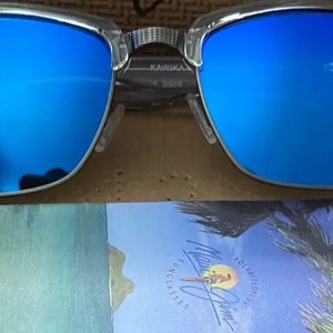 May include: Clear-framed sunglasses with vibrant blue lenses. The sunglasses have a silver metal frame and a classic design. The lenses reflect a bright blue hue. The packaging below the sunglasses features the brand name and a tropical scene.