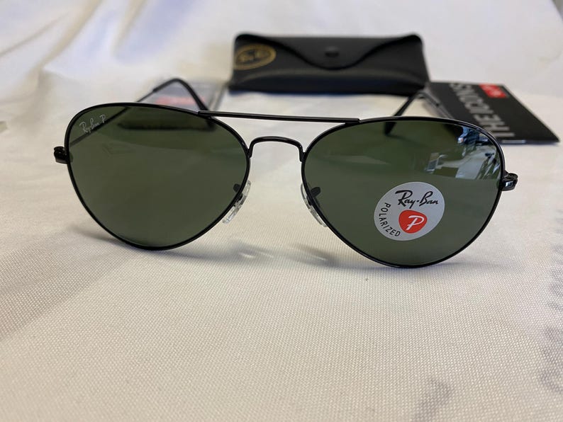 以下が含まれることがあります： 黒いRay-Banサングラス、レンズはグリーンです。サングラスはクラシックなアビエーター型で、フレームは黒です。レンズにはRay-Banのロゴが見えます。黒いケースと小冊子も画像にあります。