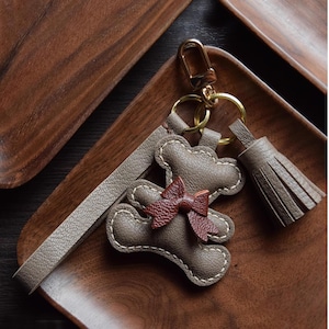 K&ouml;nnte beinhalten: Leder-Schl&uuml;sselanh&auml;nger mit Teddyb&auml;r-Design und kleiner Schleife. Der Schl&uuml;sselanh&auml;nger ist taupefarben mit wei&szlig;en N&auml;hten und einer dunkelbraunen Schleife. Er hat einen goldfarbenen Verschluss und eine passende Quaste.