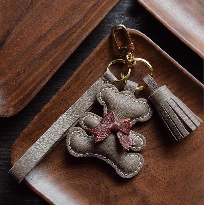 K&ouml;nnte beinhalten: Leder-Schl&uuml;sselanh&auml;nger mit Teddyb&auml;r-Design und kleiner Schleife. Der Schl&uuml;sselanh&auml;nger ist taupefarben mit wei&szlig;en N&auml;hten und einer dunkelbraunen Schleife. Er hat einen goldfarbenen Verschluss und eine passende Quaste.