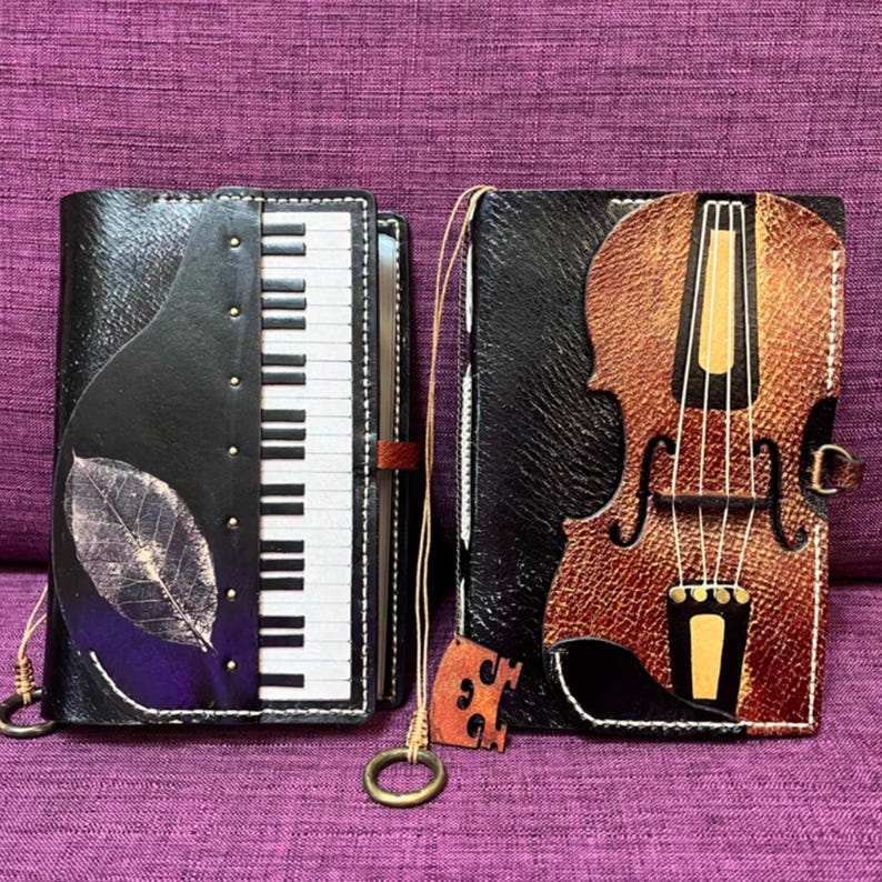 Pu&ograve; includere: Due diari in pelle con disegni di strumenti musicali. Uno presenta una tastiera di pianoforte e una foglia, l'altro un violino. Entrambi hanno copertine nere, cuciture decorative e una chiusura. I diari sono su una superficie viola.