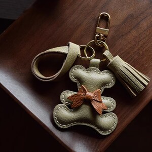 K&ouml;nnte beinhalten: Ein handgefertigter Leder-Schl&uuml;sselanh&auml;nger in Form eines Teddyb&auml;ren. Der B&auml;r ist olivgr&uuml;n mit einer braunen Schleife. Er hat einen goldfarbenen Verschluss und eine passende olivgr&uuml;ne Quaste. Der Schl&uuml;sselanh&auml;nger liegt auf einer dunkelbraunen Holzoberfl&auml;che.