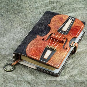 May include: A brown leather journal with a cutout design of a violin. The journal has a black and brown leather cover with a stitched edge.