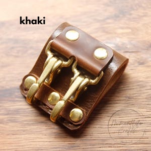 May include: Khaki leather cuff with brass hardware. The cuff features two brass hooks and is secured with brass rivets. The word "khaki" is printed in the upper left corner. The cuff is designed to wrap around the wrist.