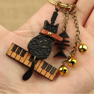 May include: A black cat-shaped keychain with a brown leather collar, a piano key design, and a fishbone charm. The keychain also features three small gold bells and a gold-colored keyring.
