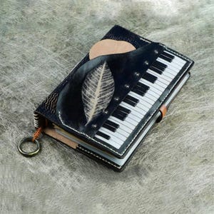 Pu&ograve; includere: Un diario in pelle fatto a mano con un design a tastiera di pianoforte. La copertina &egrave; nera con un design a foglia e sole. Il diario ha un cinturino in pelle marrone e un anello di metallo attaccato sul lato.