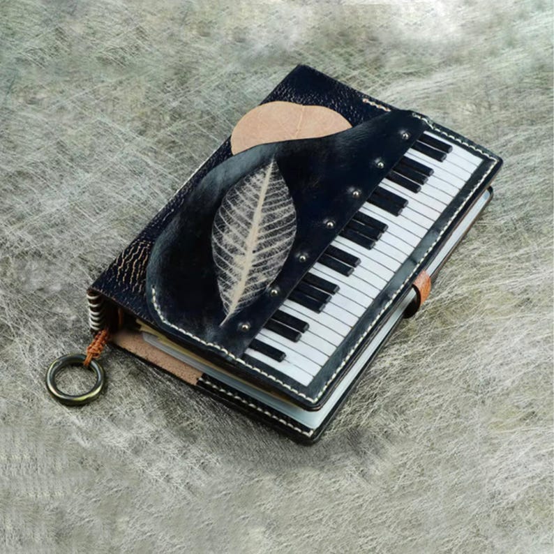 Pu&ograve; includere: Un diario in pelle fatto a mano con un design a tastiera di pianoforte. La copertina &egrave; nera con un design a foglia e sole. Il diario ha un cinturino in pelle marrone e un anello di metallo attaccato sul lato.