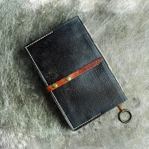 Pu&ograve; includere: Portafoglio in pelle nera con cinturino in pelle marrone fissato da due rivetti color ottone. Il portafoglio ha cuciture bianche sui bordi e un anello di metallo attaccato. La pelle ha un aspetto strutturato.