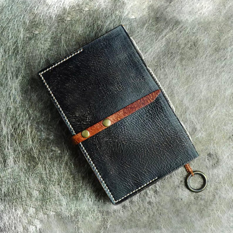 Pu&ograve; includere: Portafoglio in pelle nera con cinturino in pelle marrone fissato da due rivetti color ottone. Il portafoglio ha cuciture bianche sui bordi e un anello di metallo attaccato. La pelle ha un aspetto strutturato.