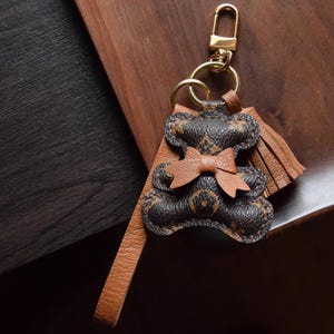K&ouml;nnte beinhalten: Ein brauner und schwarzer Leder-Teddyb&auml;r-Schl&uuml;sselanh&auml;nger mit einem goldfarbenen Verschluss und Ring. Der B&auml;r hat eine Schleife und eine Quaste. Der Schl&uuml;sselanh&auml;nger liegt auf einer Holzoberfl&auml;che.