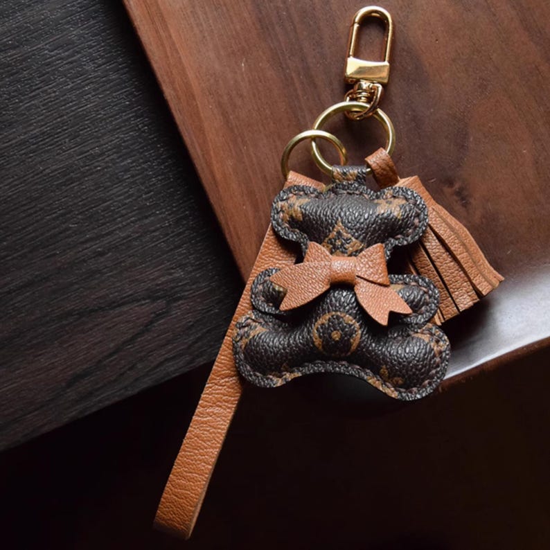K&ouml;nnte beinhalten: Ein brauner und schwarzer Leder-Teddyb&auml;r-Schl&uuml;sselanh&auml;nger mit einem goldfarbenen Verschluss und Ring. Der B&auml;r hat eine Schleife und eine Quaste. Der Schl&uuml;sselanh&auml;nger liegt auf einer Holzoberfl&auml;che.