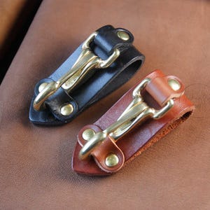 May include: Two leather keychains with brass hooks. One keychain is black leather and the other is brown leather.