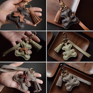 K&ouml;nnte beinhalten: Leder-Teddyb&auml;r-Schl&uuml;sselanh&auml;nger in verschiedenen Farben, darunter Braun, Olivgr&uuml;n und Taupe. Jeder Schl&uuml;sselanh&auml;nger hat eine Schleife, eine Quaste und goldfarbene Hardware. Die Schl&uuml;sselanh&auml;nger sind mit einer gen&auml;hten Kontur versehen.