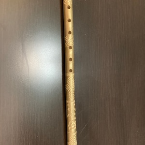 May include: A brass flute with intricate floral and geometric patterns etched along its length. The instrument has several finger holes and a polished finish, reflecting the light. The flute is a musical instrument.