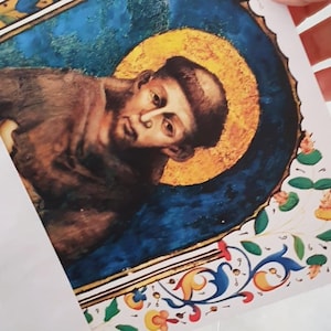 May include: A print of a religious figure with a golden halo, set against a blue background. The artwork features ornate floral and scrollwork borders in blue, yellow, and red. The figure is depicted in brown robes.
