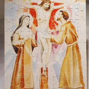 May include: A completed jigsaw puzzle depicting a religious scene. The image shows a figure on a cross with two other figures in robes. The puzzle pieces are interlocked, forming a complete picture with a light background.