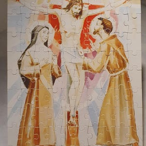 May include: A completed jigsaw puzzle depicting the crucifixion of Jesus Christ. The artwork features Jesus on the cross, with figures in robes and a vibrant sky background. The puzzle is displayed on a metal stand.